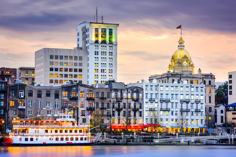 Historic Savannah riverfront buildings and a paddlewheel riverboat at sunset.